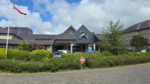 The outside of Noble's Hospital, which has a three-legs above the entrance and an area of greenery with a no entry sign in front of it.