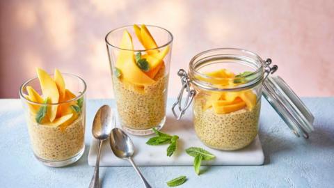 Mango lassi chia seed pudding in a mason jar and two glasses. There are two tea spoons on a board.