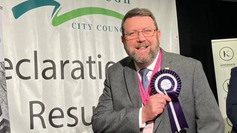 Chris Harper with short grey hair, a short beard and glasses. He is wearing a grey suit, white shirt and a blue tie, and is pointing to a round purple rosette pinned to his suit jacket following an election. He is smiling at the camera.