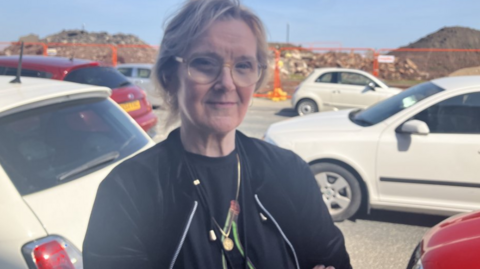 Karen Webb-Meek - a woman with short blonde hair wearing glasses and a black jacket over a black T-shirt. She is standing by a busy road with lots of cars behind her. Next to the road are piles of rubble and soil. The sky is blue with no clouds.