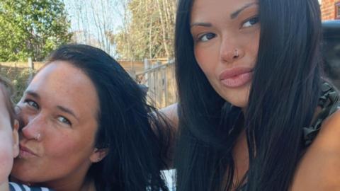 Image of two women with long black hair and nose rings smiling at the camera. In the background can be seen a garden fence, trees and plants.