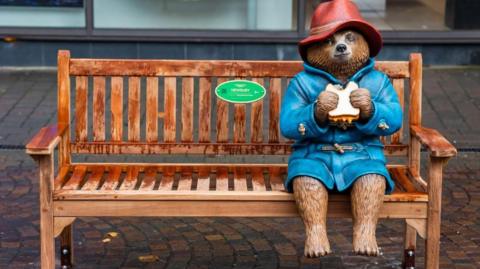 A statue of a Paddington Bear in a red hat and a blue coat holding a sandwich and sitting on a bench 