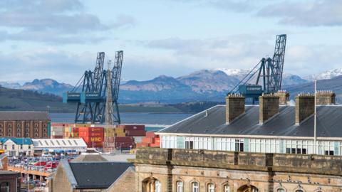A yard full of shipping containers next to a river with snow-capped hills on the faraway bank