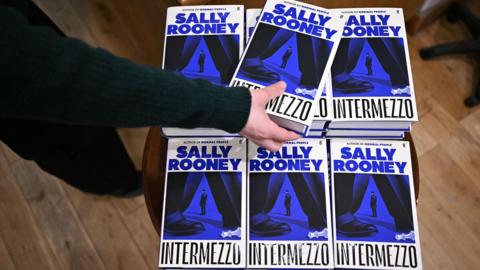 A person's hand picks up a copy of Intermezzo - the new book by Sally Rooney - from a pile of books