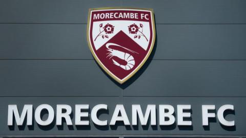The Morecambe Football Club crest above the name 'Morecambe FC', which is written in capital letters, as seen on the front of their home ground, the Mazuma Mobile Stadium