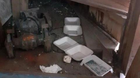 Three white takeaway food containers on a dark, dirty floor. They have black specks inside them. A rusty piece of equipment in on the floor close to the containers. There is also a scrunched up paper napkin on the floor.