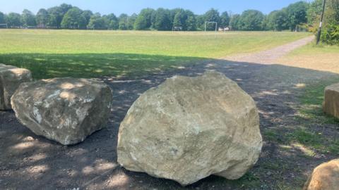 Boulders in front of a grassy playing field surrounded by trees on a bright sunny day.