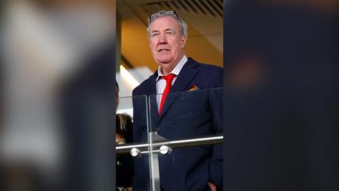 Jeremy Clarkson is wearing a navy suit, red tie, and white shirt with a black grid pattern.