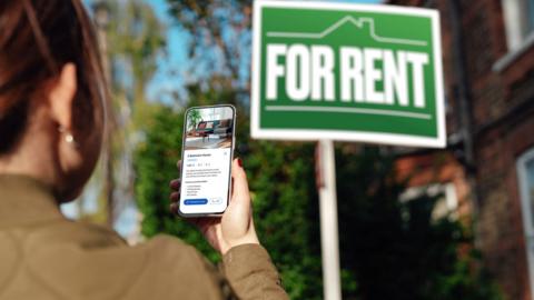 A woman in front of a green sign which says 'For Rent' in white letters. She is looking for information about the house on her phone and is wearing a beige jacket and has brown hair tied back. 