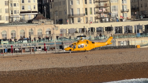 A helicopter on a pebbled beach with cream Regency-style buildings in the background