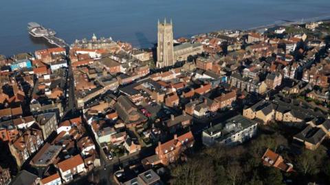An aerial of Cromer in Norfolk, overlooking the seaside town towards the coast. The photograph features a range of properties and buildings including Cromer Parish Church (St Peter and St Paul) which is a tall building towering above the town and Cromer Pier which leads out to the sea. 
