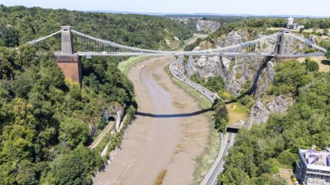 Drone view of A4 Portway running along under the Clifton Suspension Bridge and the River Avon. It's a bright sunny day and there's lots of green vegetation on either side of the river. The river is running dry and above skies are light blue.