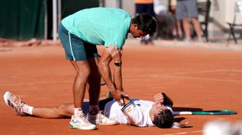 Jakub Mensik lies on the floor and opponent Mariano Navone tries to assist him