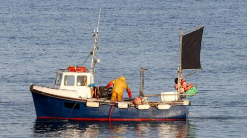 A fishing boat in the sea. A fisherman, wearing yellow overalls, handles a lobster pot as they stand on the deck of a white and blue coloured vessel.