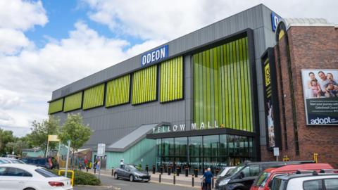 The Metrocentre's Yellow Mall . A glass entrance stands at the front of the building. Yellow slats run up the side of the building and a sign advertises a cinema inside. Car are parked in the car park.