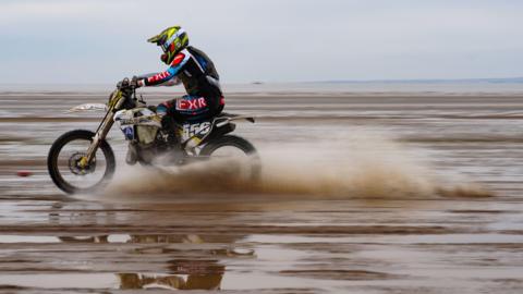 A person rides a white dirt bike with the number 556 along weston beach. It is muddy with water being thrown up by the bike. The rider wears a black red and blue kit and yellow and black helmet