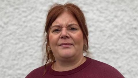 The image shows Michelle Brace, a woman with red hair and a burgundy top standing with her back to a white pebble-dash wall.