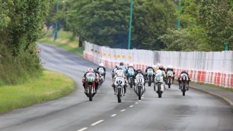 A group of motorcycles riding up the course at Billown, with crash barriers and fencing to the right and greenery to the left.