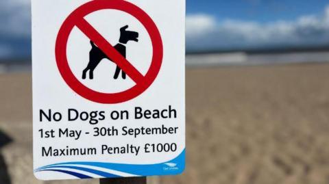 A sign on a beach saying No Dogs on Beach from May 1 to September 30. Maximum penalty £1,000. There is a dog in a red circle with a line through it and a beach in the background.