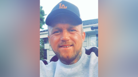 A photo of Marcus Carpenter. He is smiling at the camera, he has a beard and tattoos on his neck. He is wearing a grey sweatshirt with black details, and a black cap.