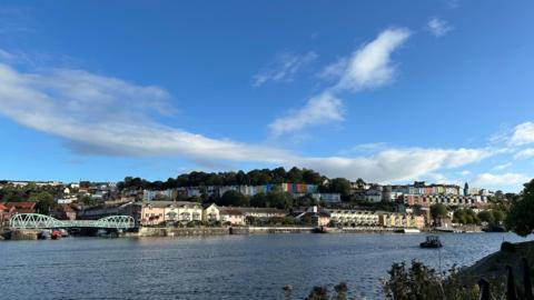 A wider river with colourful houses on the far bank under a fair weather sky