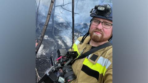 Ed Matthews, who wears a protective uniform, holds a camera. He is stood in a smoky woodland area.