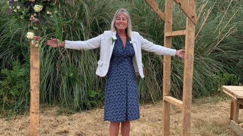 A woman holding her arms out, stands at a wooden frame decorated with flowers. She is standing outside on dry grass with greenery behind her and a table to her right.