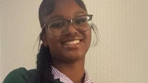 A close-up photo of a smiling Elianne Andam, she has long black hair in a braid and is wearing glasses. She is wearing a striped school uniform blouse.