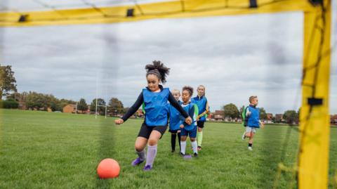 A group of children playing football on a large grassy field. The viewpoint is from behind a small yellow goal frame, looking outward toward the players. A bright orange football sits in the foreground near the opening of the goal. A child wearing a blue training bib and dark sports clothing is positioned closest to the goal, appearing to run or move toward the ball. Behind this child, several other children, also wearing blue training bibs, are spread out across the field. One child in the background is wearing a green goalkeeper-style bib and standing further back.