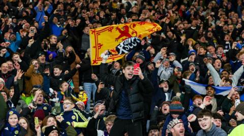 Dozens of Scotland fans celebrating at the final whistle at Hampden. They have scarves and banners.
