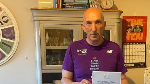 John McNally standing in his kitchen and holding a handbag appeal poster. He is wearing a purple London Marathon finisher's t-shirt.