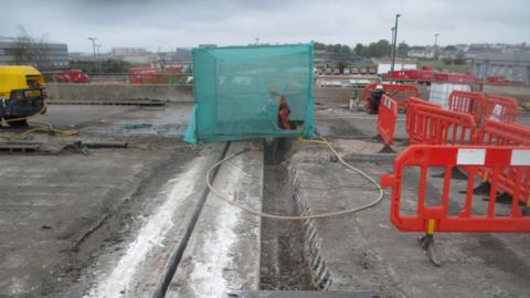 A worker in high-vis can be seen behind a green mesh screen amid the roadworks, working on a large trench. There are a number of orange barriers across the road, and a yellow digger to the left.