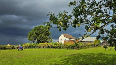 Very dark grey clouds above a white house with a horse in a field below 