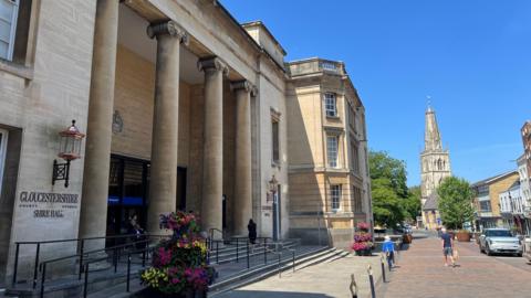An external street level view of Gloucestershire Shire Hall, with flower baskets placed outside the front of the building. The picture is taken on a sunny, bright day