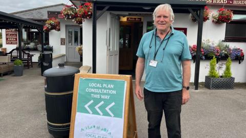 Adrian Birch, Green Party leader, wearing a green shirt. He is smiling and standing outside a pub, next to a wooden A-board with "local plan consultation this way" written on it.