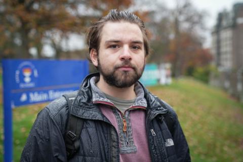 Fraser Adams has short, brown hair and a beard. He is standing outdoors on a cloudy day, wearing a rain-speckled black jacket and backpack. A blue sign and autumn trees are visible in the background.