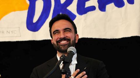 New York mayoral candidate, State Rep. Zohran Mamdani in front of a mic with hand on his heart.