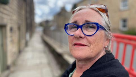 Older woman wearing blue-framed glass on a street in front of orange crash barriers