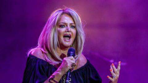 Bonnie Tyler on stage in purple pink lighting wearing black top and holding microphone. She has long blonde hair worn down.