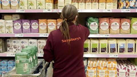 A supermarket shop worker stacks shelves with flower, on the back of her uniform the Saisnbury's log can be seen