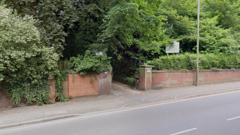 A view of from the other side of a road showing a brick wall with a path leading to a nursing home. There are many large trees with green foliage hanging down above the wall. There is a white sign protruding from the bushes.