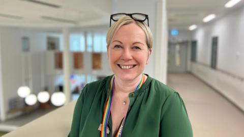 Laura Skaife-Knight standing in a corridor, wearing a green shirt. She has a rainbow lanyard round her neck. She's smiling and has blonde hair, with her glasses on her head. 