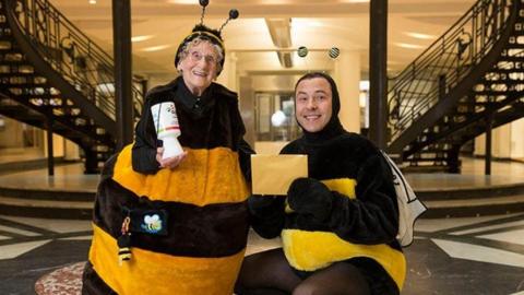 Jean Bishop dressed in her bee outfit holding a collecting tin, with actor David Walliams also dressed as a bee. He is kneeling down and holding an envelope.