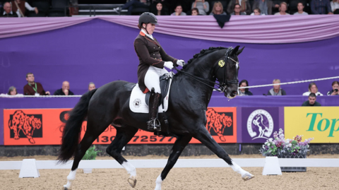 A woman riding a horse inside an arena doing dressage. The horse is black with white socks and a white flash on its forehead. The woman is wearing a black helmet with a brown jacket, white trousers and black boots