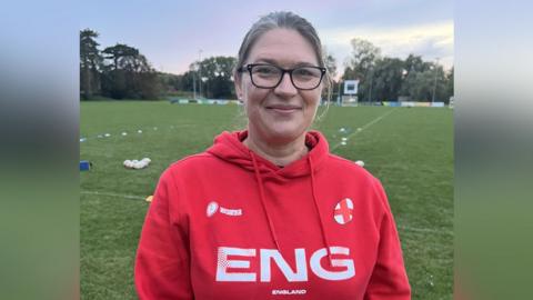 Helen Durman in a red England rugby hoody. Her hair is tied back and she is wearing glasses. Behind her is a rugby pitch with training equipment laid out. 