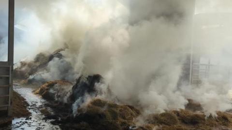 Thick smoke rises from a pile of smouldering hay spread out on the floor of a large barn