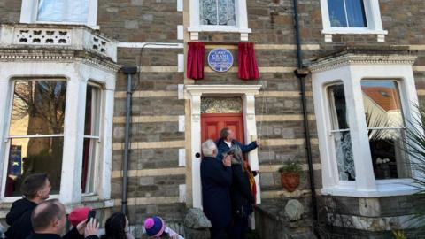 Three people stand underneath a blue plaque beneath a red curtain.