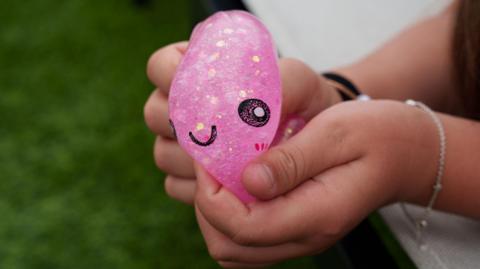 Child’s hands squeezing a pink glitter squishy dumpling toy. The toy has a smiley face on it. The child is wearing a silver bracelet.