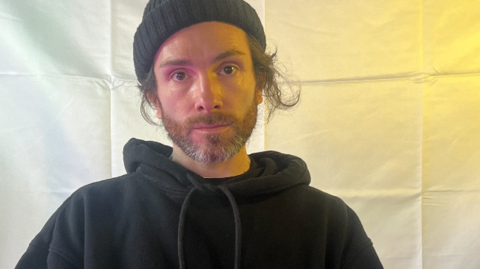 A man wearing a black hoodie and a black hat is sitting in front of a white sheet that is lit up from behind. 