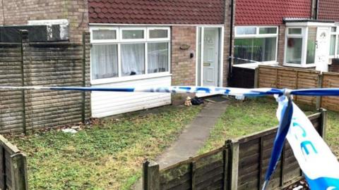 Police tape, in front of a house, with the tape going across the image from left to right. There is grass in front of the house. 